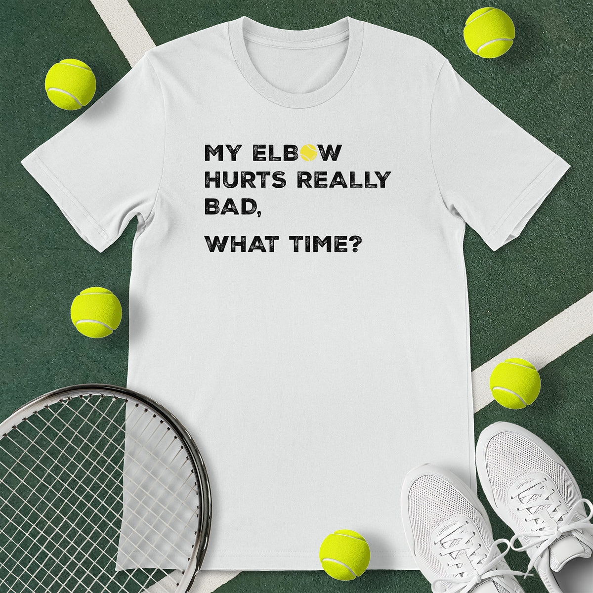 White t-shirt with the text 'MY ELBOW HURTS REALLY BAD, WHAT TIME?' and a graphic of a tennis ball.